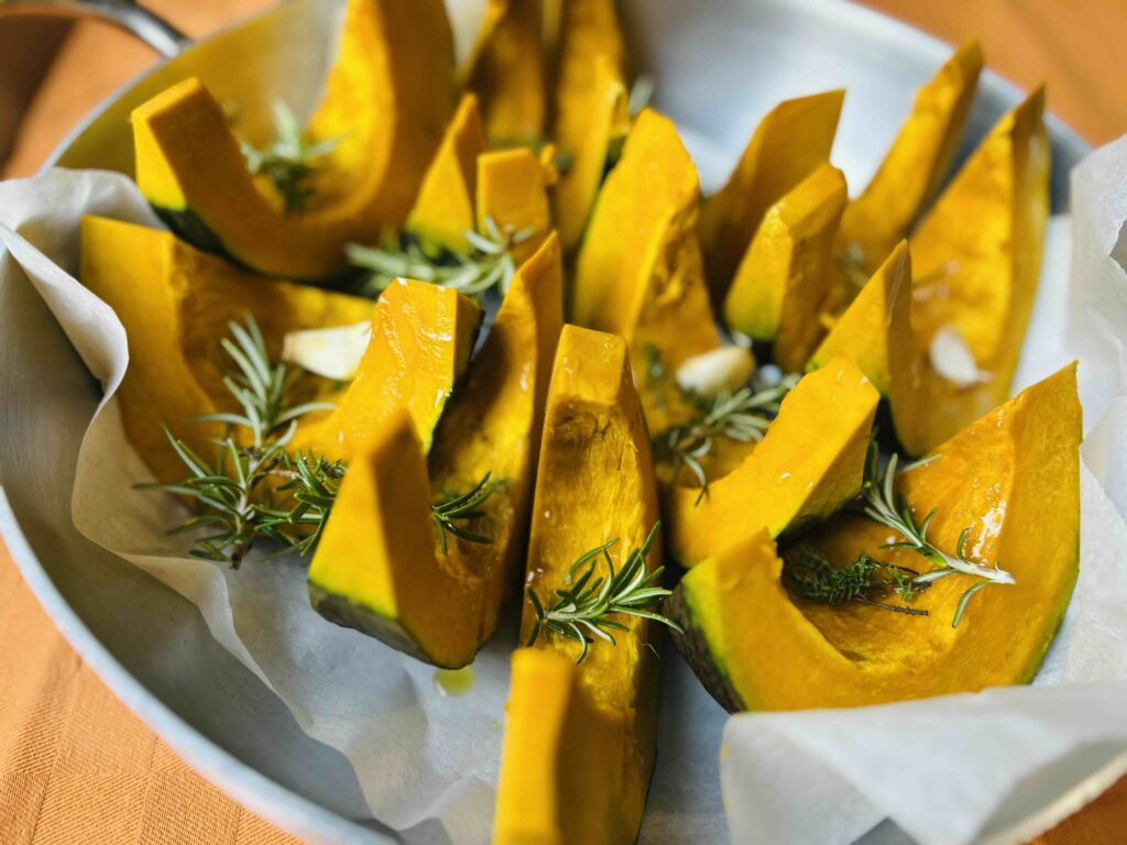 Spicchi di zucca cruda con rosmarino e aglio disposti in teglia, pronti per la cottura al forno.
